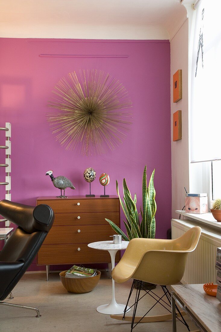 Wohnzimmer im Retrostil mit einer lilafarbenen Wand, Kunstobjekten und einem Eames Schaukelstuhl