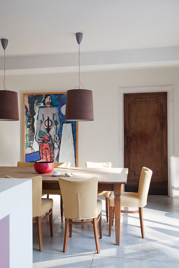 Rustic dining table and chairs with cream upholstery in front of modern painting and wooden door