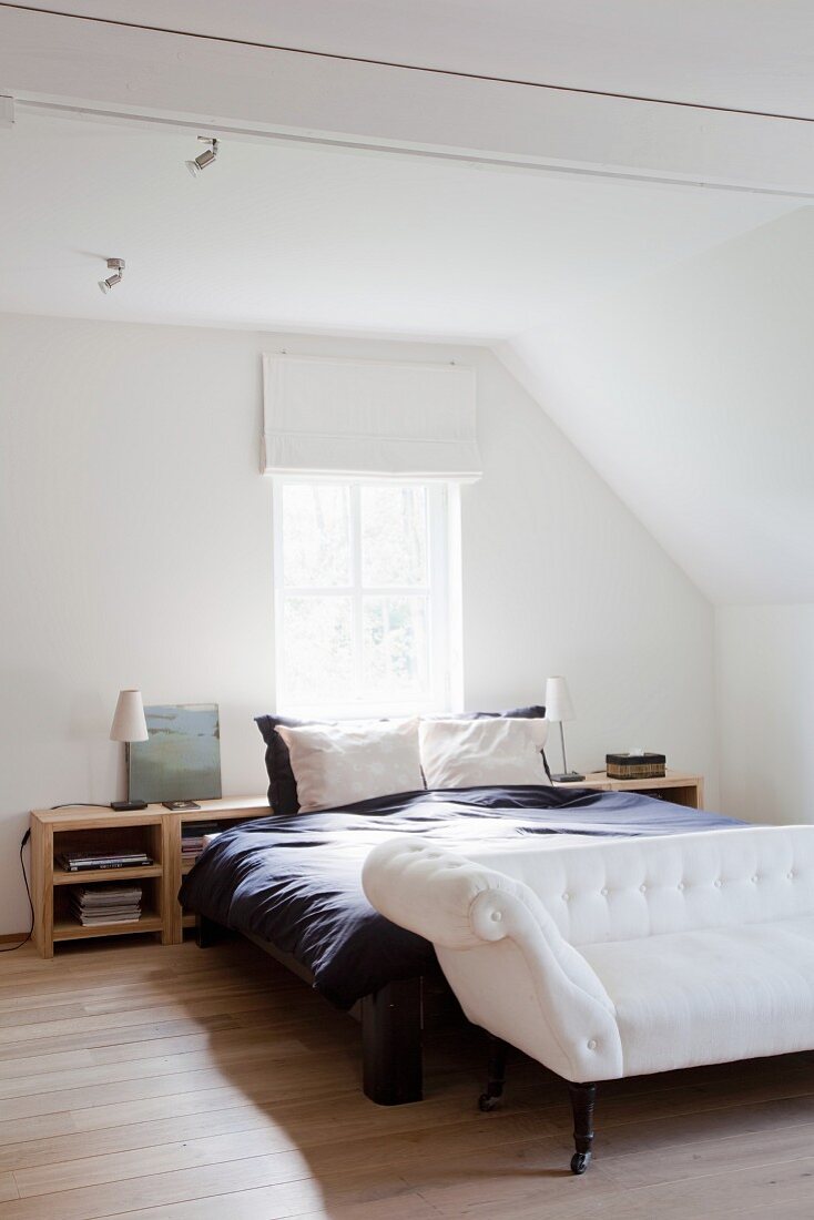 Elegant, antique-style white sofa at foot of double bed in white attic bedroom with oak parquet floor