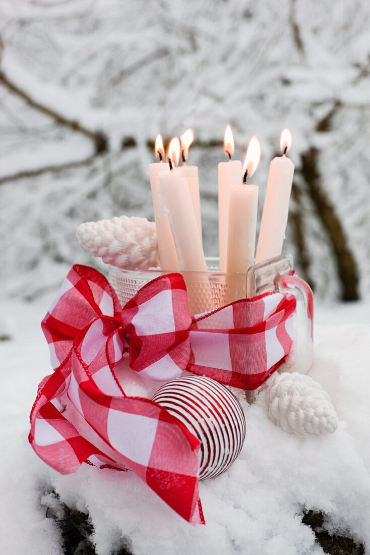 Lit candles in old glass scoop decorated with red ribbon and white Christmas baubles surrounded by snow