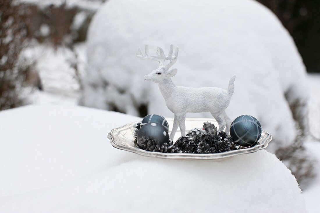 weiße Deko-Hirschfigur mit Metall Tannenzapfen und Christbaumkugeln auf Teller im Schnee
