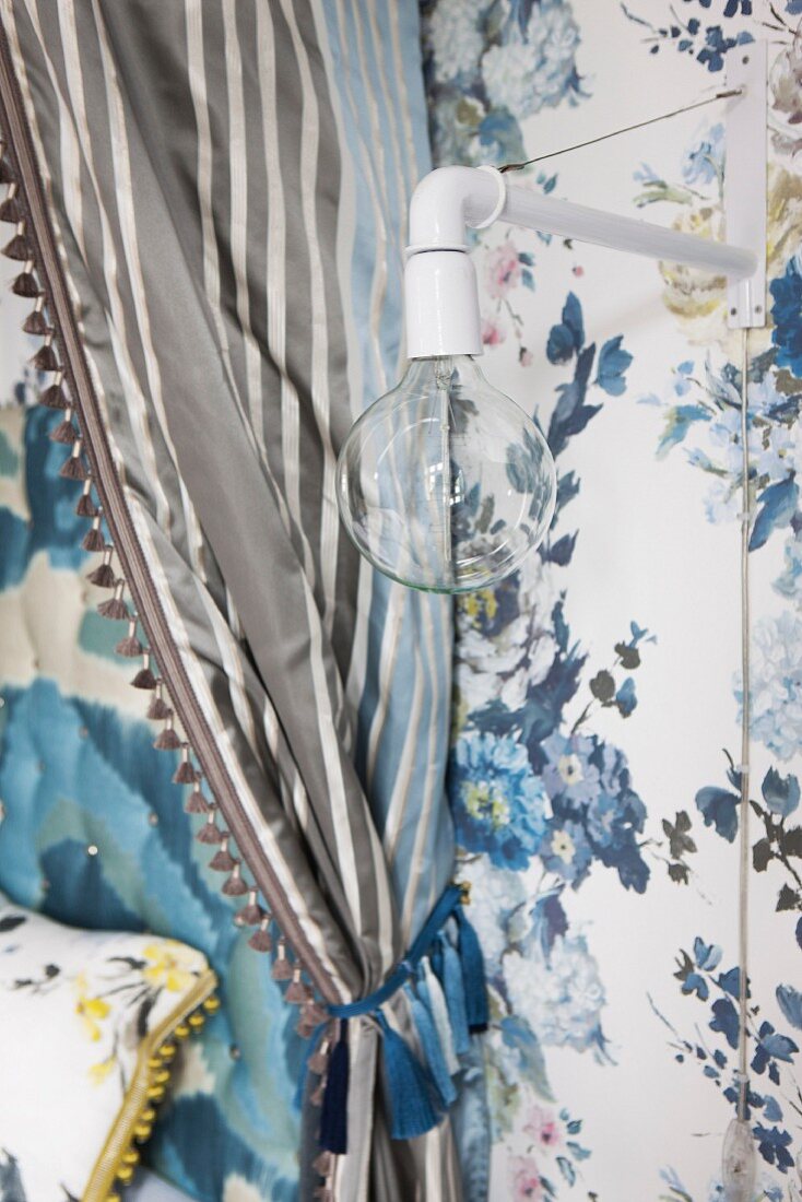 Minimalistische Wandleuchte an tapezierter Wand mit blauem Blumenmuster neben drapiertem Baldachin-Vorhang