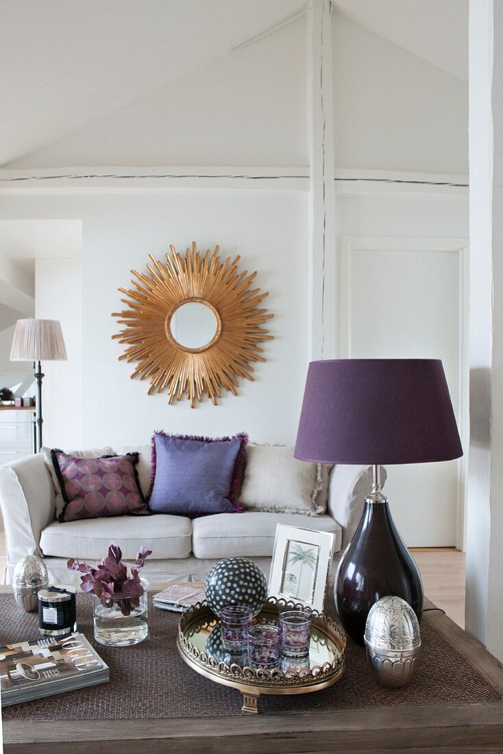 Couchtisch mit violetter Tischleuchte dahinter Sofa und sonnenförmiger Wandspiegel