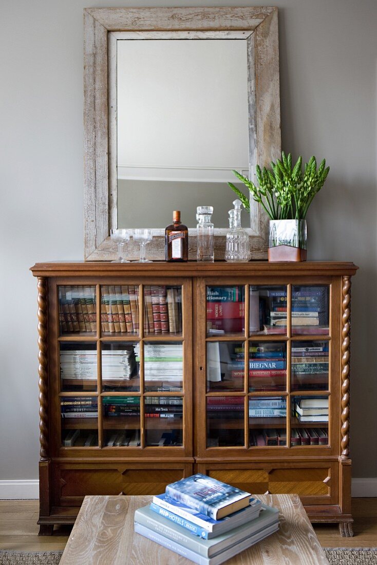 Mirror leaning against pale grey wall on top of antique, half-height, glass-fronted cabinet