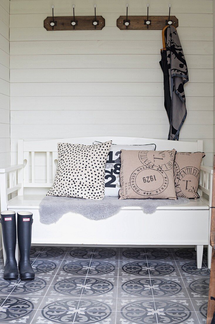 Weiß lackierte Holzbank mit Kissen vor weisser Holzwand oberhalb Hakenleiste, auf Boden Fliesen mit Retro Muster
