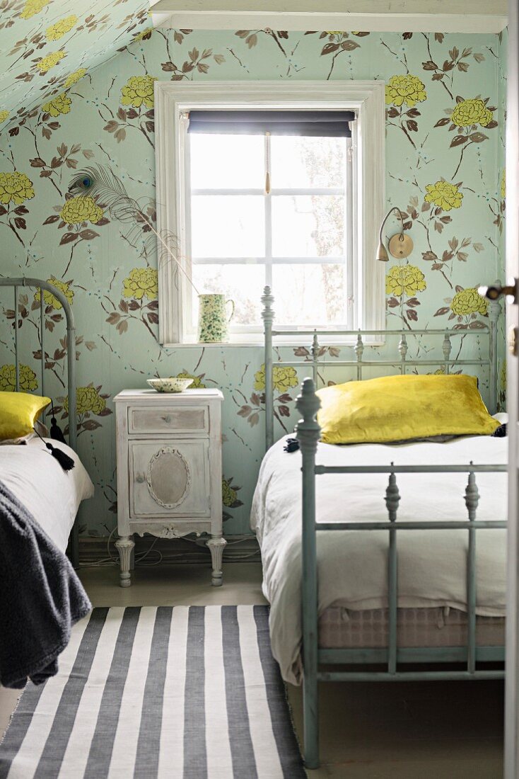 Vintage-style bedroom with floral wallpaper and bedside cabinet between twin metal beds with frames painted grey below window