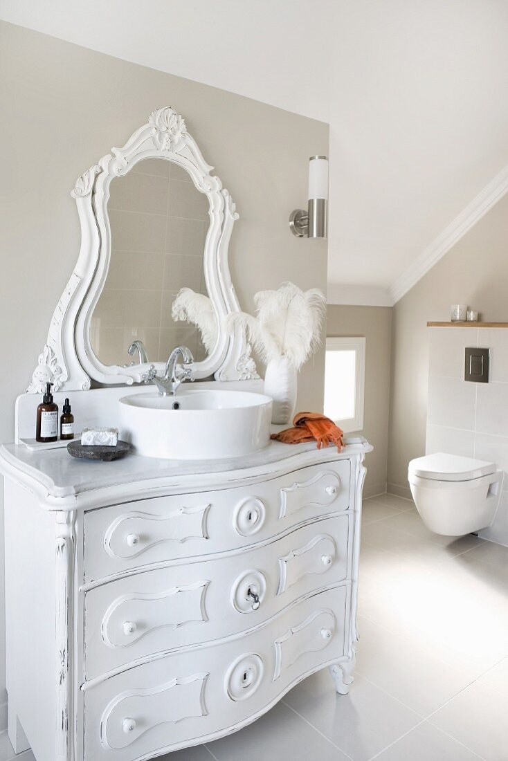 Modern bathroom with sloping ceiling and wall-mounted toilet; vintage chest of drawers painted white with round, modern countertop washbasin and carved mirror