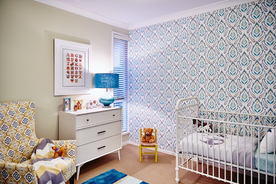 White cot against retro wallpaper and patterned armchair next to white chest of drawers