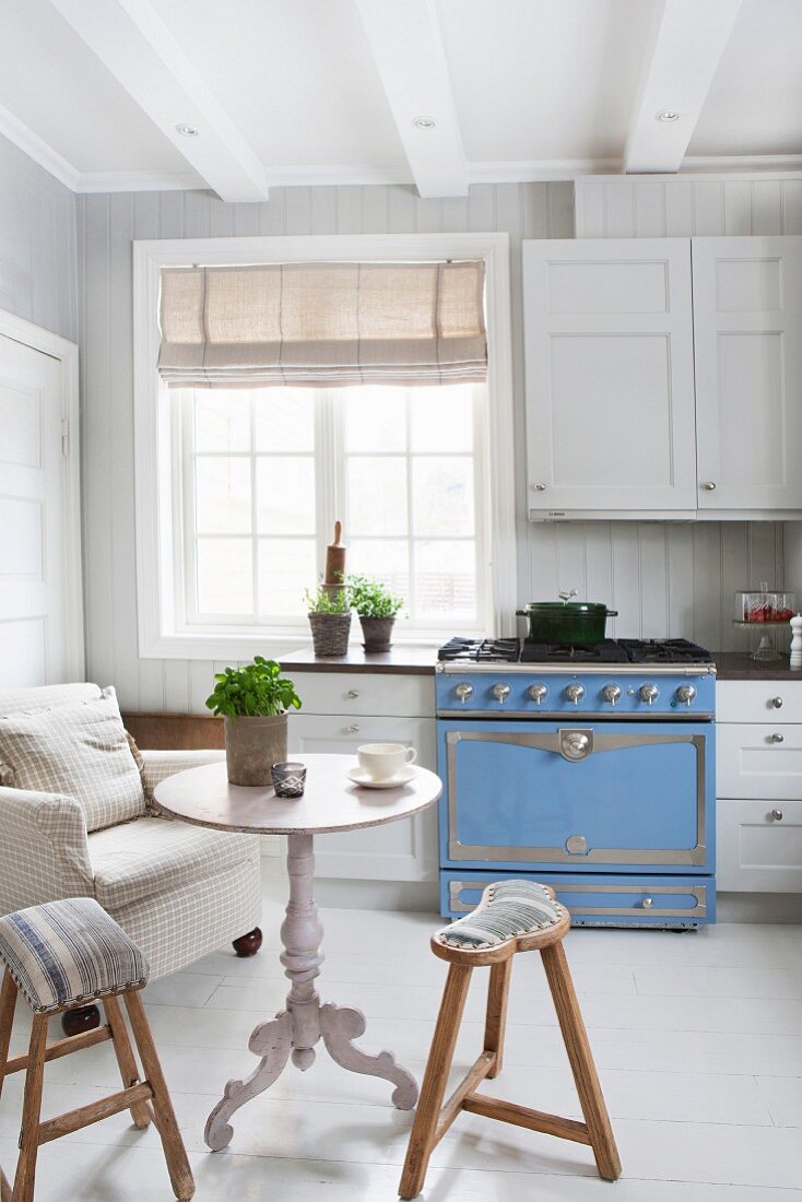 weiße, skandinavische Landhausküche mit hellblauem Gasherd, Polstersessel, Tisch und Holzhocker mit nostalgischem Flair