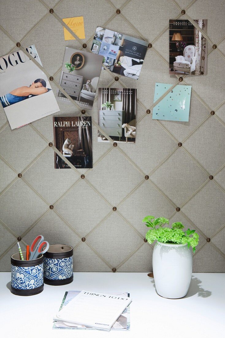 Postcards stuck on elegant fabric-covered pinboard above pen holders and vase of flowers on white desk