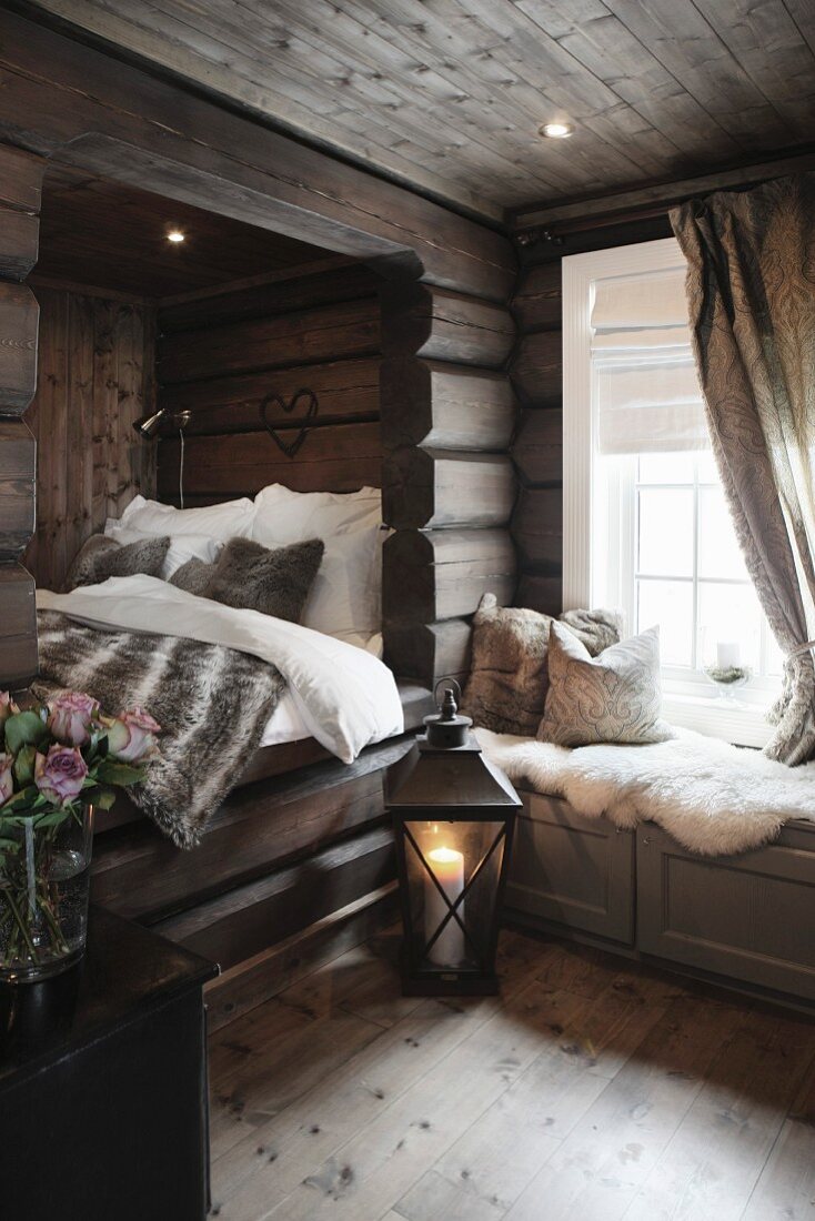 Bodenlaterne mit brennender Kerze neben Alkoven, Fell Tagesdecke auf Bett, seitlich eingebaute Sitzbank vor Fenster in Holzhaus