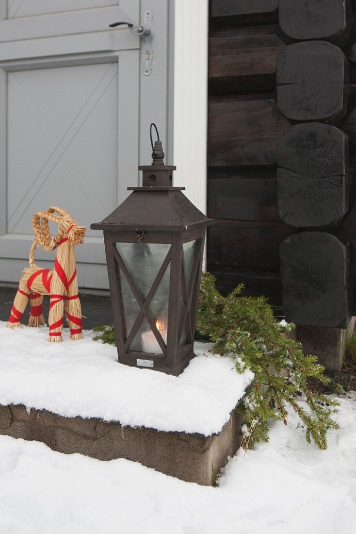 Tierfigur aus Stroh neben Bodenlaterne mit brennender Kerze auf verschneiter Stufe