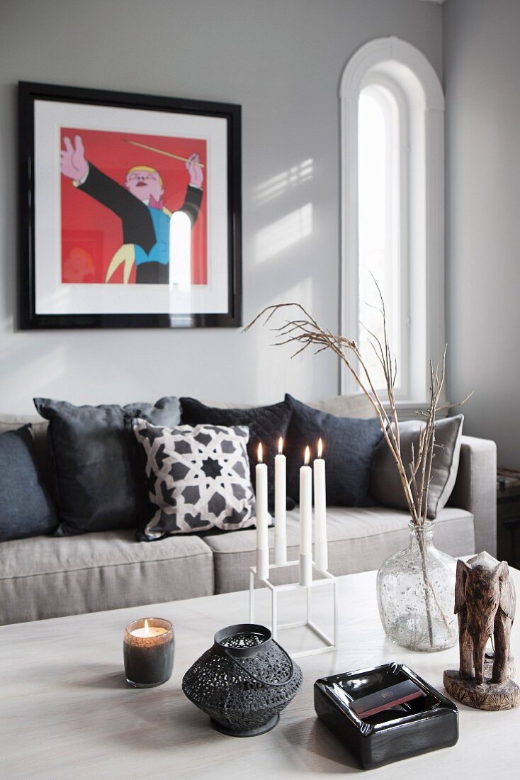 Lit candles in candelabra and tealight holder on table, scatter cushions on sofa below modern artwork on grey wall in traditional interior