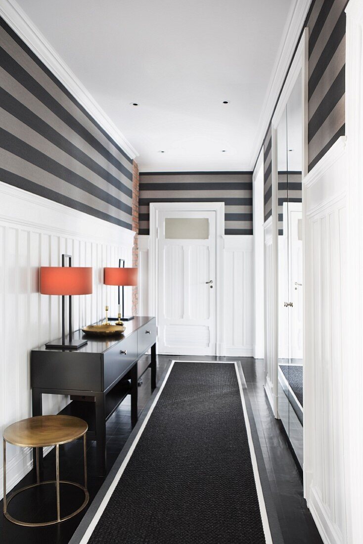 Black runner, table lamps on console table and white wainscoting below grey striped walls in elegant hallway