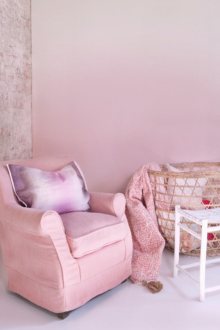 Rosa Zimmerecke, Polstersessel und Kissen neben Wäschekorb aus Geflecht vor rosa getönter Wand