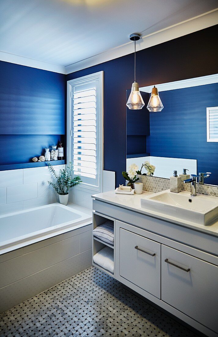 Waschtisch mit weißem Unterschrank neben eingebauter Badewanne vor Fenster in blau getöntem Bad