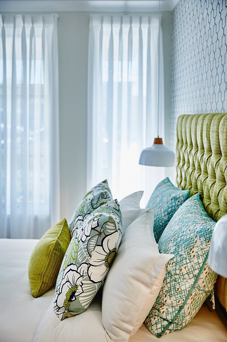 Green patterned scatter cushions on double bed with button-tufted headboard in front of white curtains