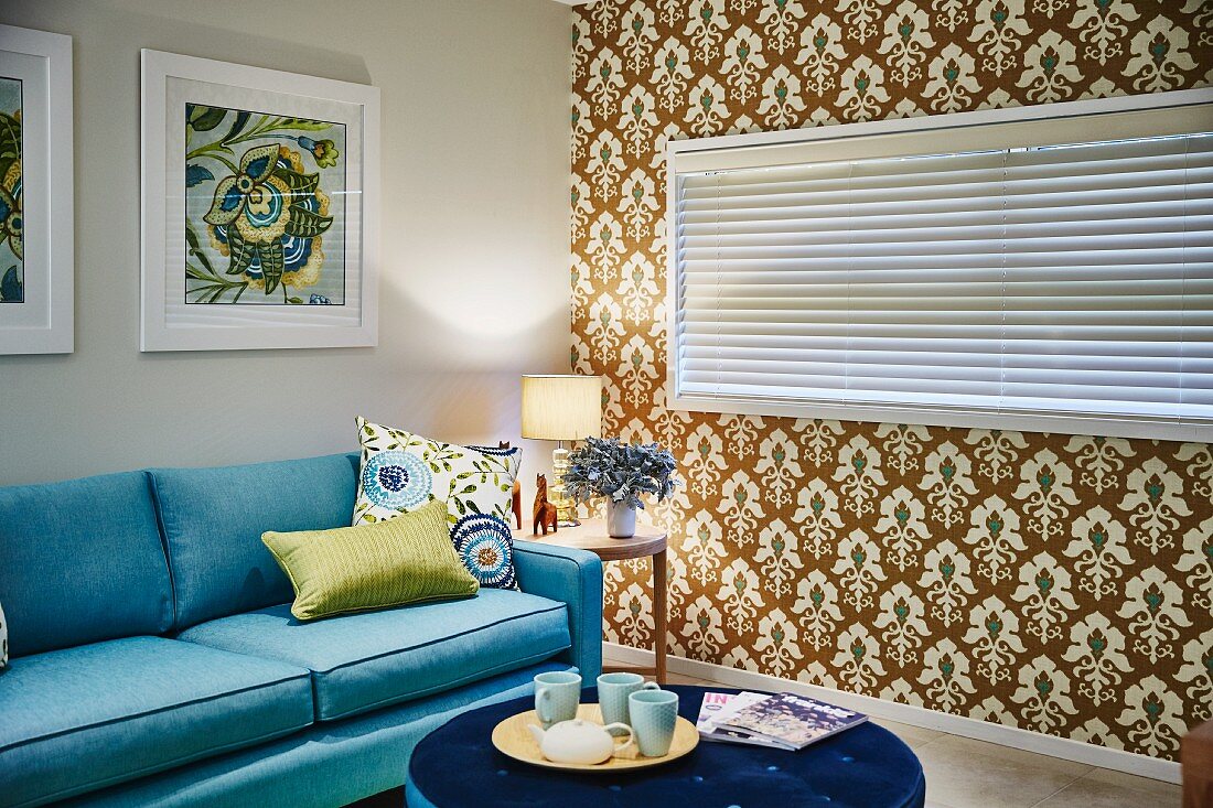 Runder Polstertisch mit Tassen auf Tablett und hellblaues Sofa in Wohnzimmer, seitlich tapezierte Wand mit Retro Ornament Muster, Fenster mit geschlossener Jalousie
