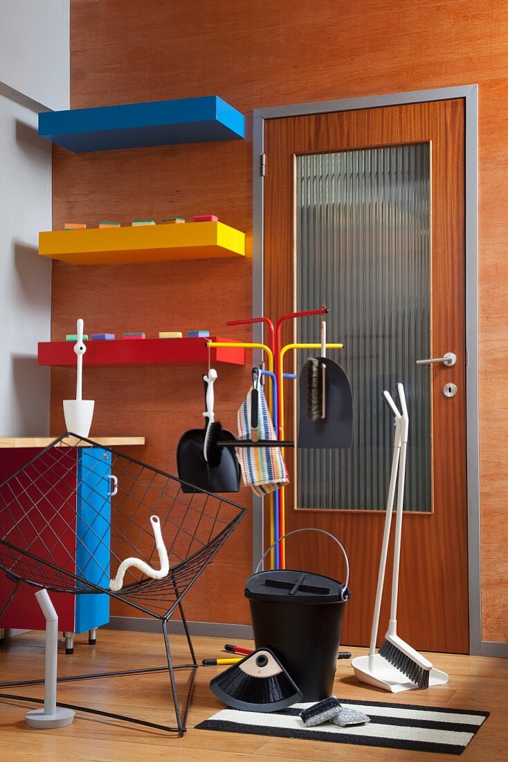 Cleaning utensils and retro wire armchair in front of brightly coloured shelved on wood-clad wall