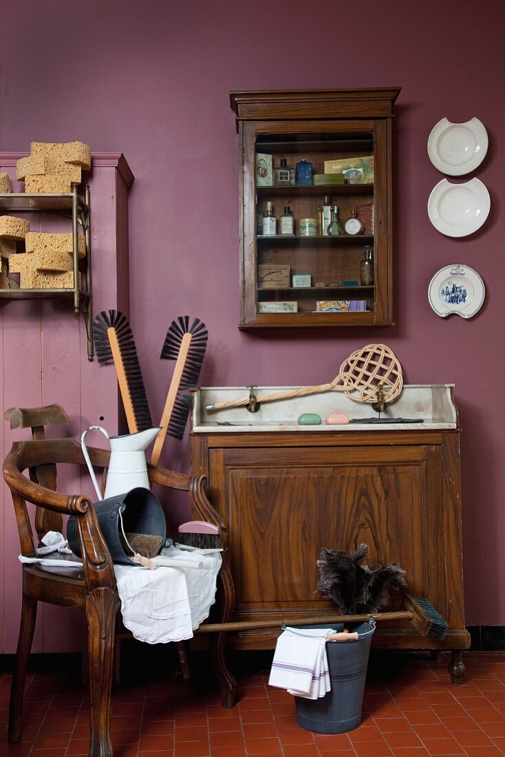 Alter Waschtisch, Wandschränkchen und antiker Armlehnstuhl mit Putzutensilien vor violetter Wand
