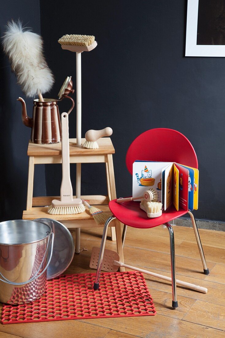 Cleaning brushes with wooden handles and feather duster on step stool; metal bucket on non-slip mat and children's book on red chair