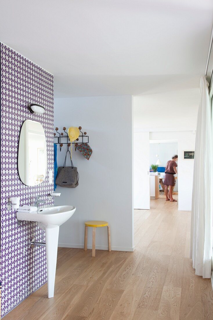 Standwaschbecken und Spiegel an tapezierter Wand mit Retro Muster in offenem Raum, Blick durch mehrere Durchgänge auf Frau im Hintergrund