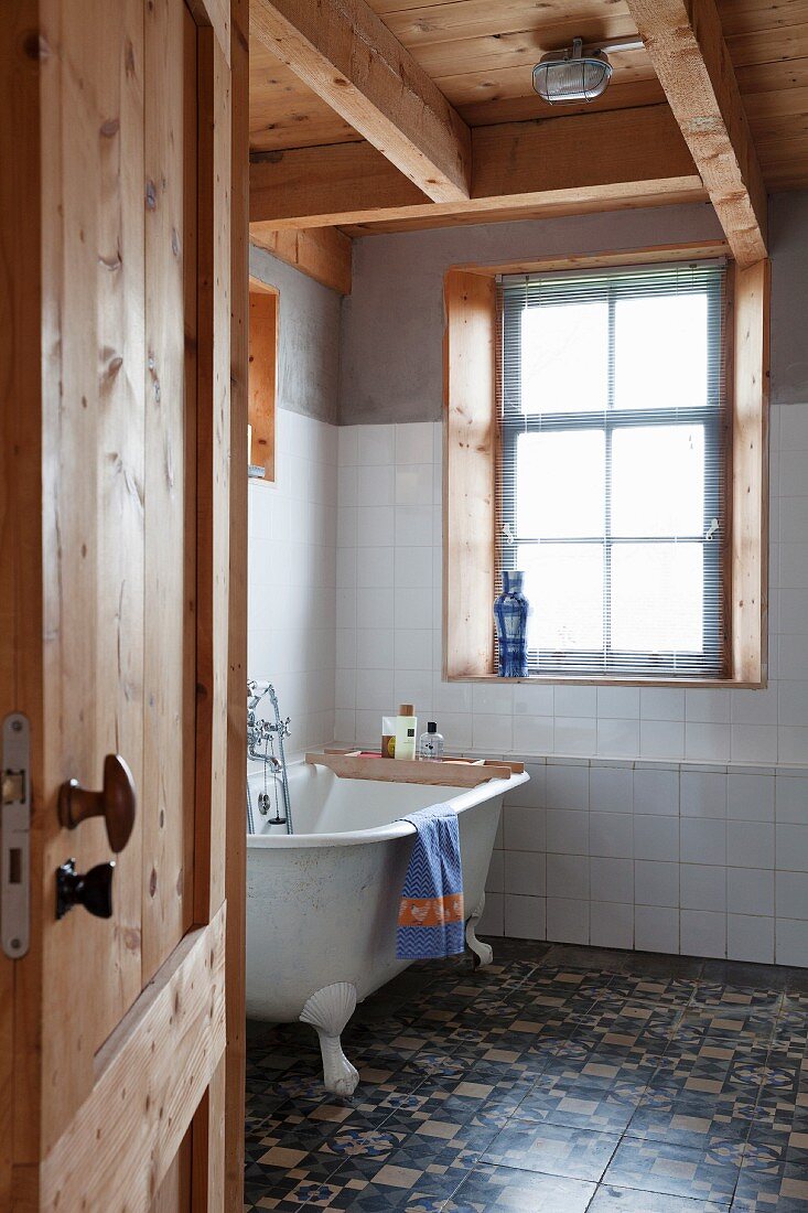 View of vintage bathtub and patterned floor tiles in rustic bathroom seen through open door