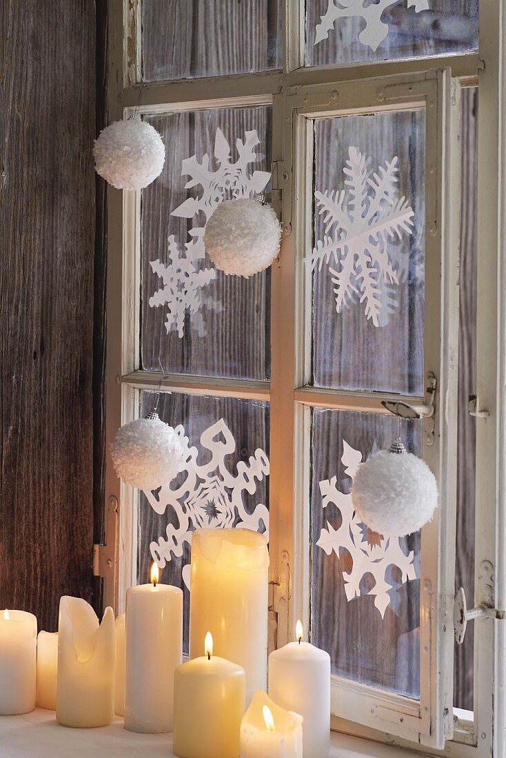 Fenster mit winterlichen Papiersternen dekoriert, davor weiße, brennende Stumpenkerzen