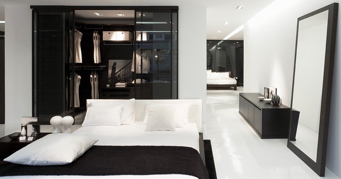 Doppelbett mit weisser Bettwäsche vor Ankleide, seitlich Standspiegel neben schwarzem Lowboard vor Wand
