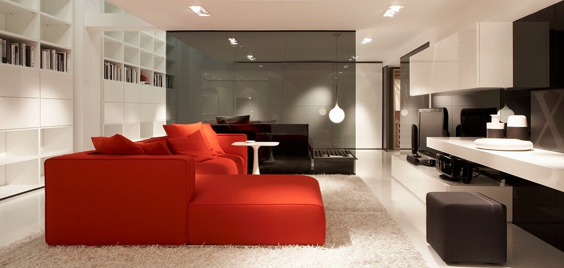 Rote Sofakombination und zierlicher Beistelltisch auf weißem Flokati-Teppich in offener Lounge, im Hintergrund rauchgraue Glastrennwand