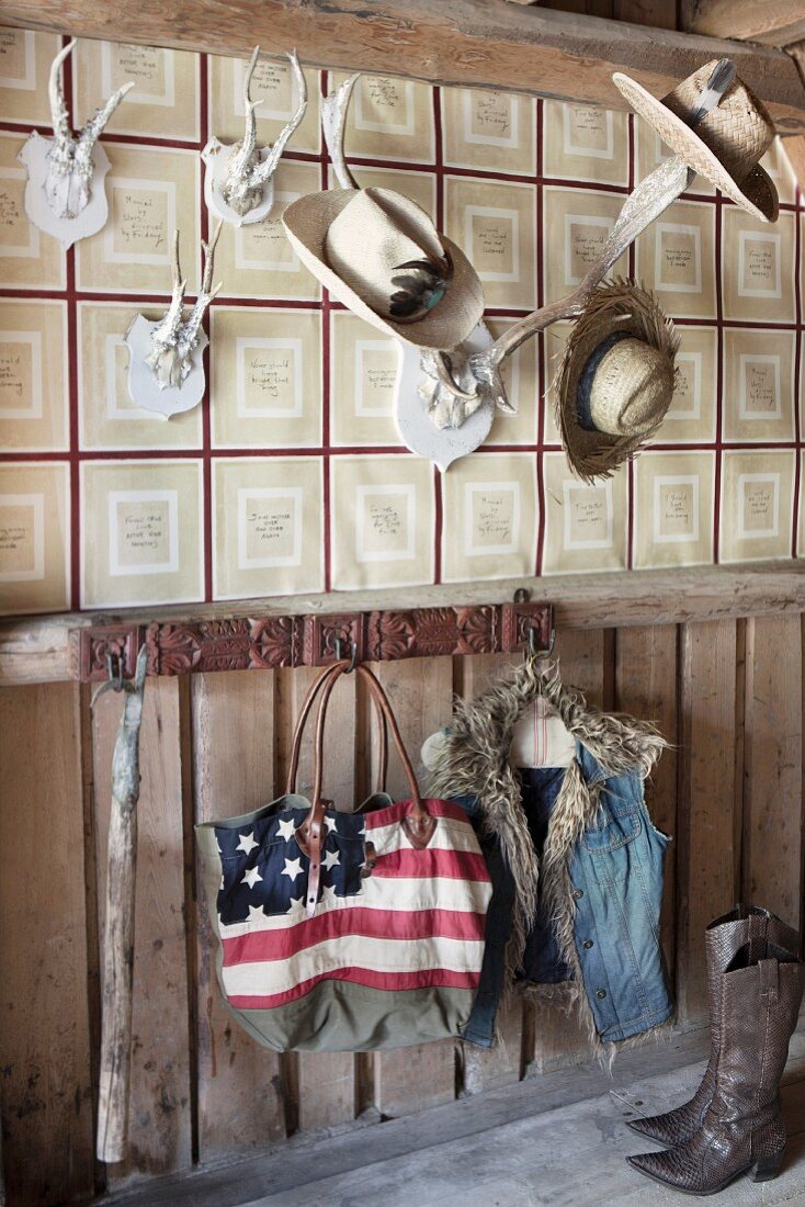 Tasche mit amerikanischer Flagge und Weste auf Wandhaken aufgehängt, darüber Tiertrophäen mit verschiedenen Hüten an tapezierter Wand