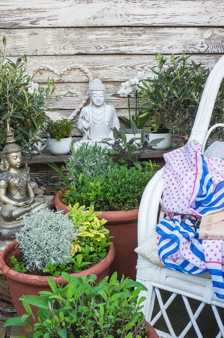 Kräutertöpfe, Buddhafiguren und weiße Orchideen neben weißem Rattanstuhl im Garten