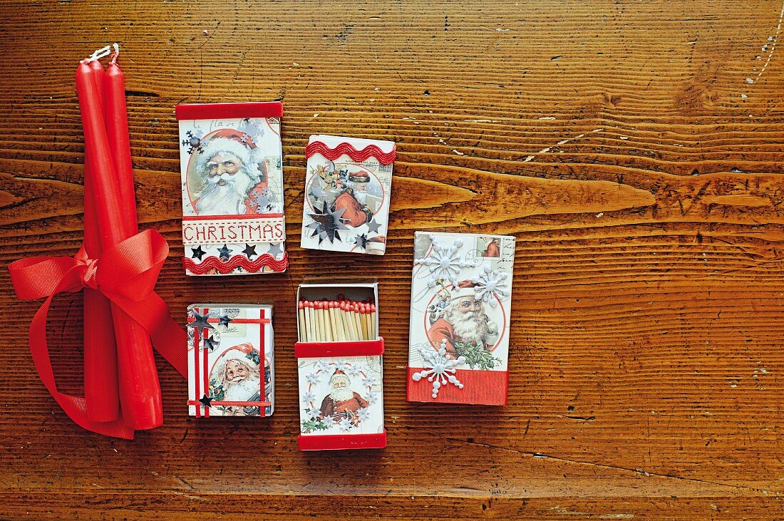 Verzierte Streichholzschachteln mit Weihnachtsmotiven beklebt und rote Kerzen mit Schleifenband auf Holzuntergrund