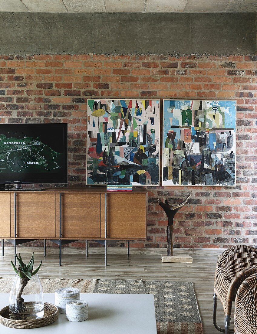 Designer-Sideboard an Ziegelwand, moderne Bilder, im Vordergrund teilweise sichtbarer, weisser Couchtisch im Wohnzimmer