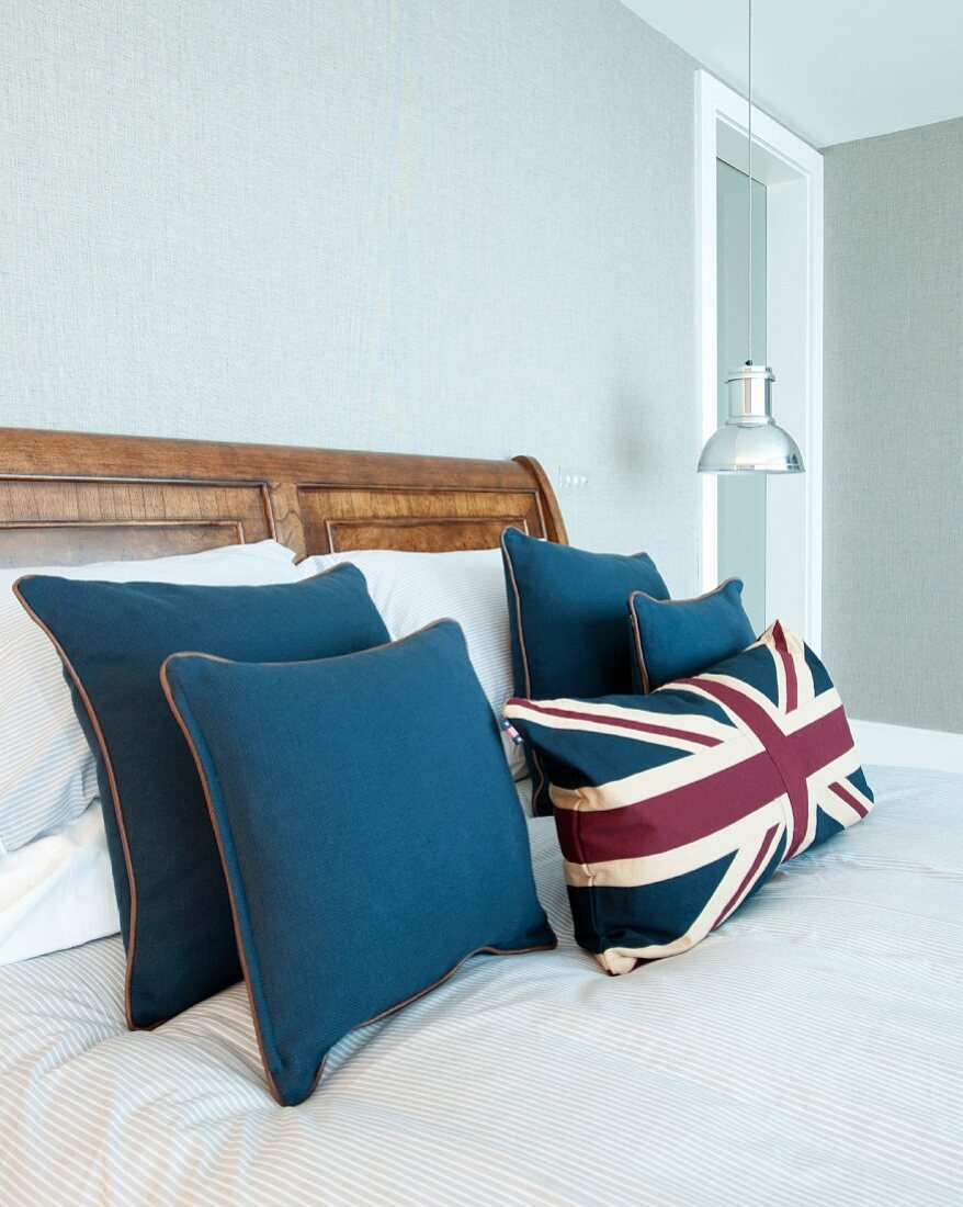 Dekokissen in Blau und mit Union Jack Motiv auf Bett aus massivem Holz; Hängeleuchte aus verchromtem Metall im Hintergrund