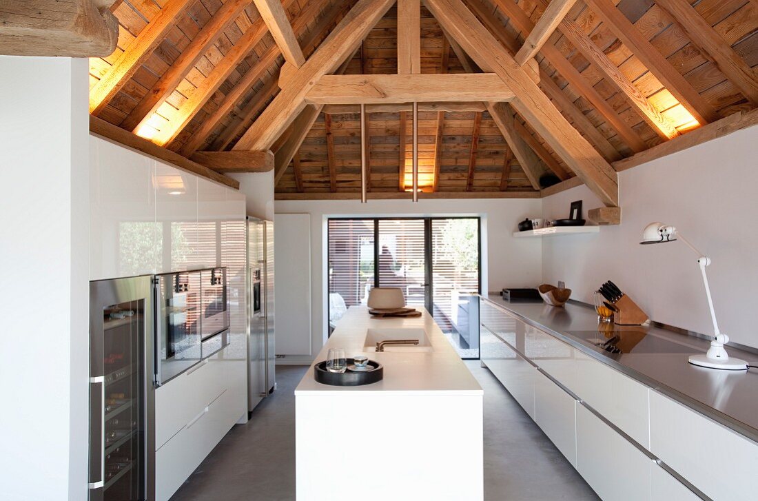 Designer kitchen with glossy white surfaces below rustic wood-beamed roof structure with indirect lighting