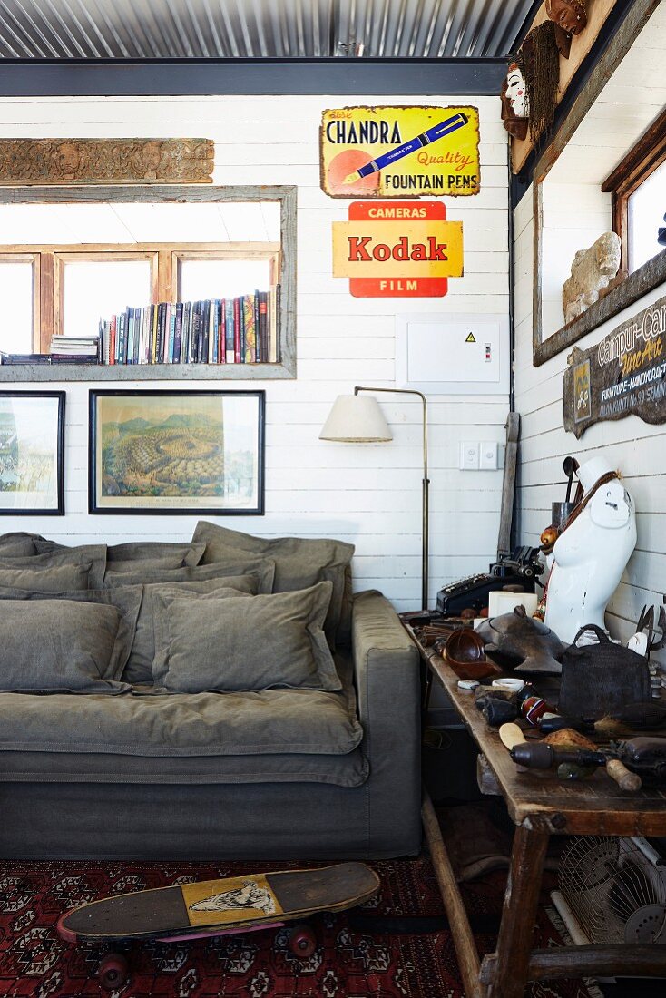 Rustikale Zimmerecke mit weisser Holzverschalung, graue Couch unter Bildern und Werbetafeln, seitlich Holztisch mit Reiseerinnerungen