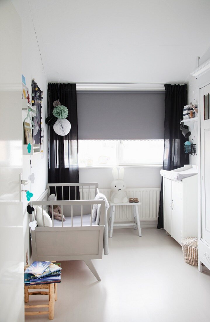 Kinderzimmer mit weißem Retro-Gitterbett und Wickelkommode, grauer Rollo und schwarze Vorhänge