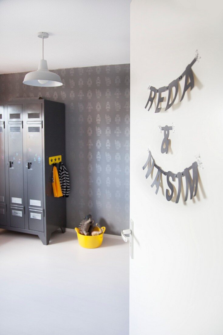 Grey-painted locker, patterned wallpaper, yellow plastic container of toys and boys names hung on door in boys' bedroom