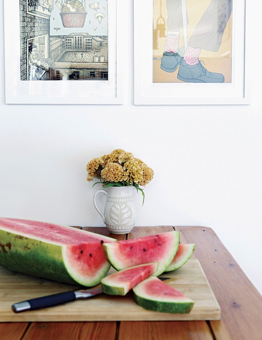 Aufgeschnittene Wassermelone auf Schneidebrett und Blumenkrug auf Tisch unter gerahmten Zeichnungen