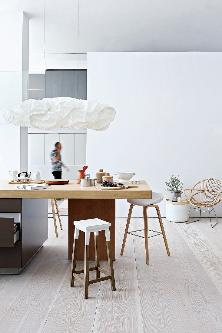 Bar stools around kitchen counter below designer pendant lamps in modern, open-plan interior with pale wooden floor