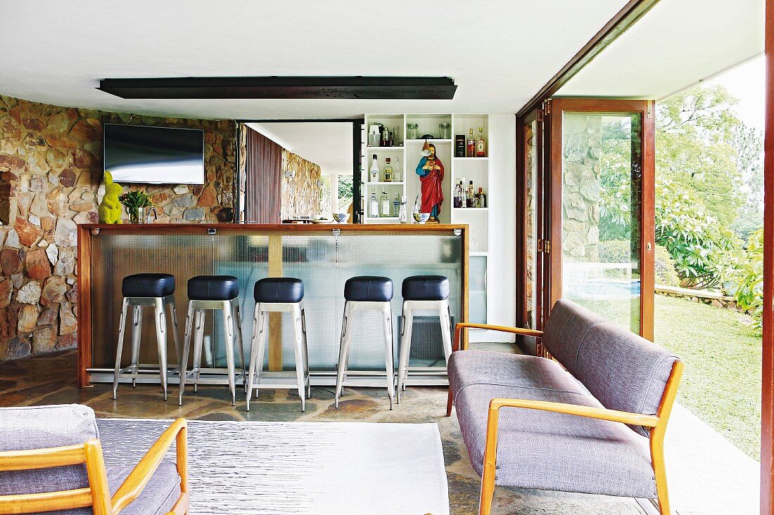 Gepolsterte Sitzbank im 50er Jahre Stil in Loungebereich, im Hintergrund Retro Barhocker vor Theke in offenem Wohnraum, seitlich Glasfalttür zum Garten