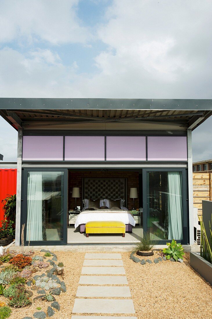 Blick von Dachterrasse mit Trittplatten in Kiesfläche durch offene Glasschiebetüren auf Schlafzimmer in Wohncontainer
