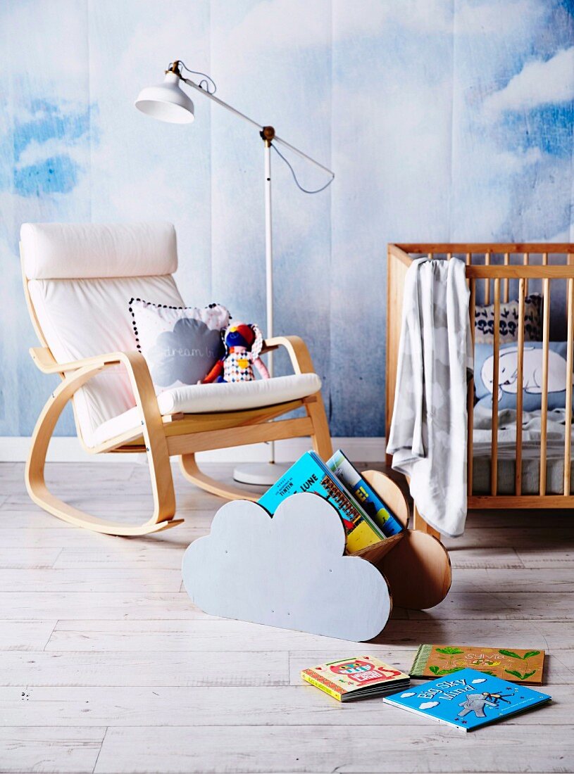Hand-crafted cloud-shaped magazine rack in front of rocking chair and standard lamp against wall painted with mural of cloudy sky