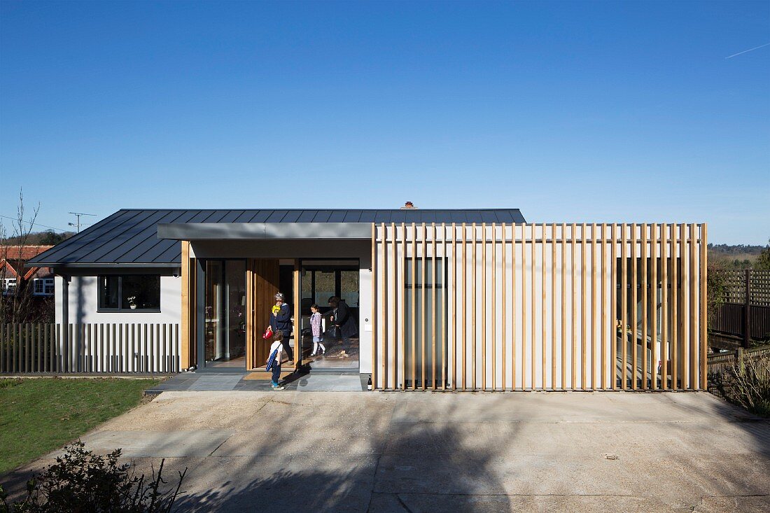 Blue sky above modern house with family in open front door and vertical wooden slat structure covering facade in rural area