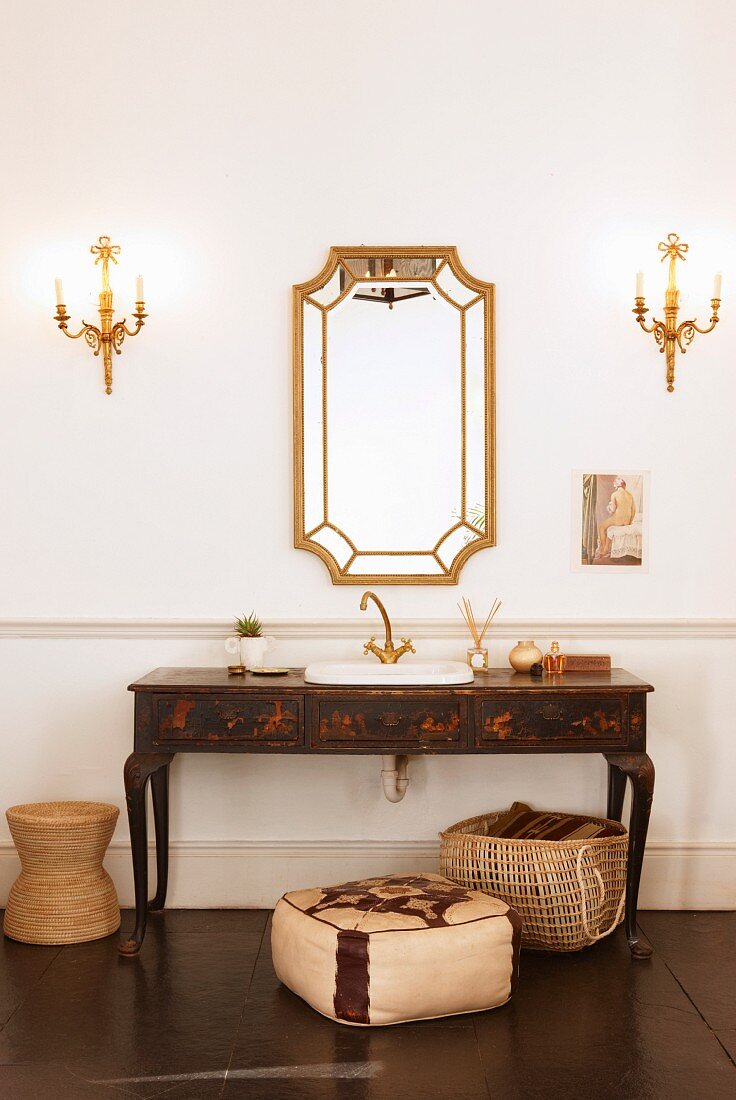 Antique, painted washstand and framed mirror flanked by candle sconces above pouffe and basket on dark floor