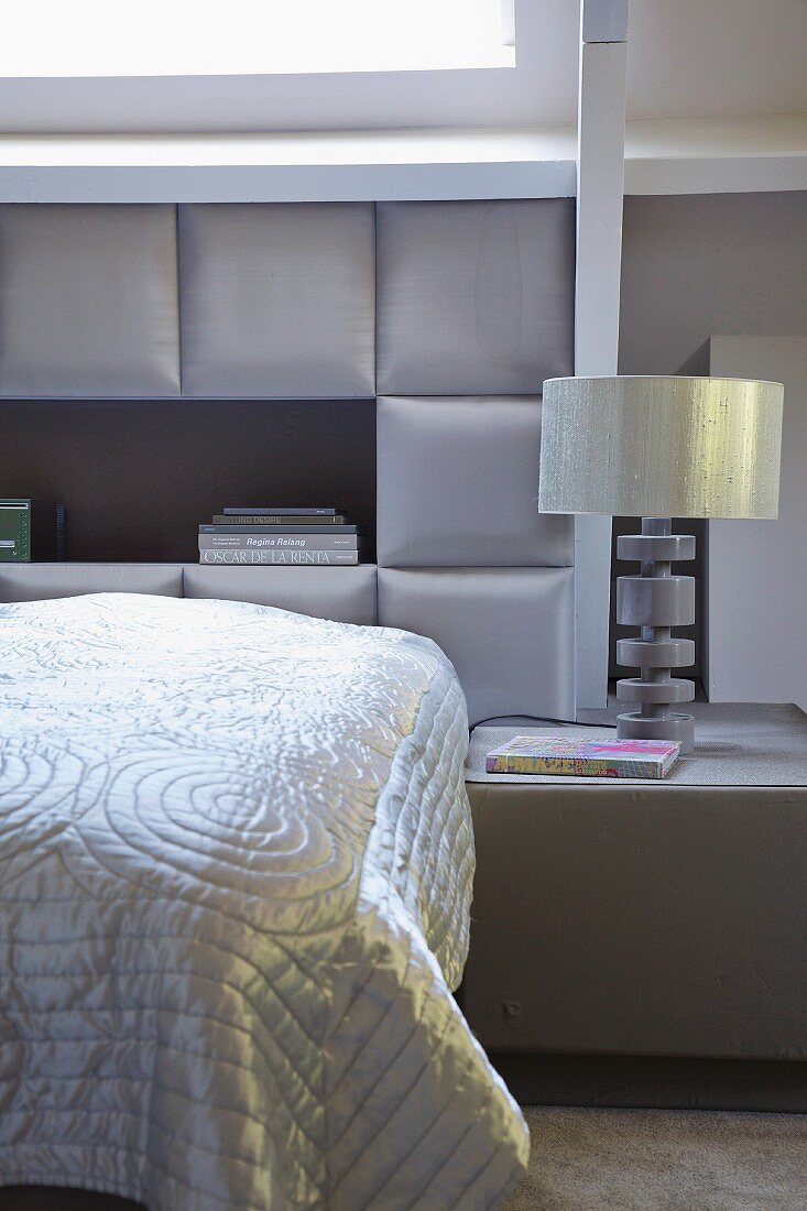 Schlafzimmer in eleganten Grautönen, auf Bett glänzende Tagesdecke vor gepolsterter Kassettenverkleidung an Wand, seitlich Nachttisch mit Tischleuchte