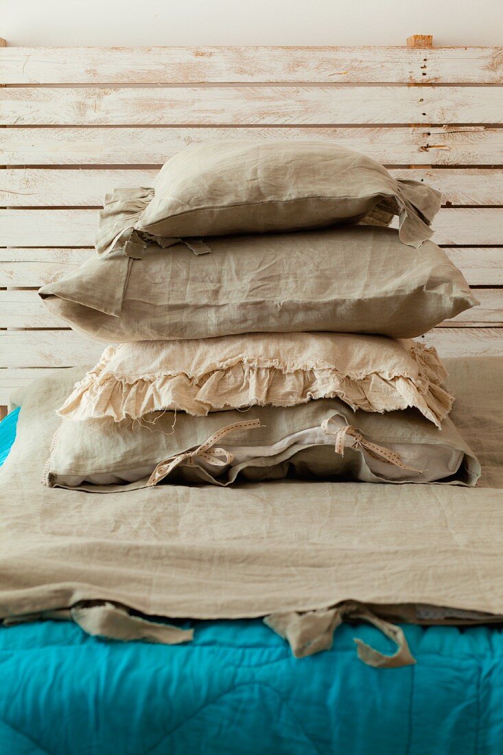 Gestapelte Kissen mit Leinenbezug auf Bett mit türkisfarbener Steppdecke und rustikalem Kopfende aus Holz