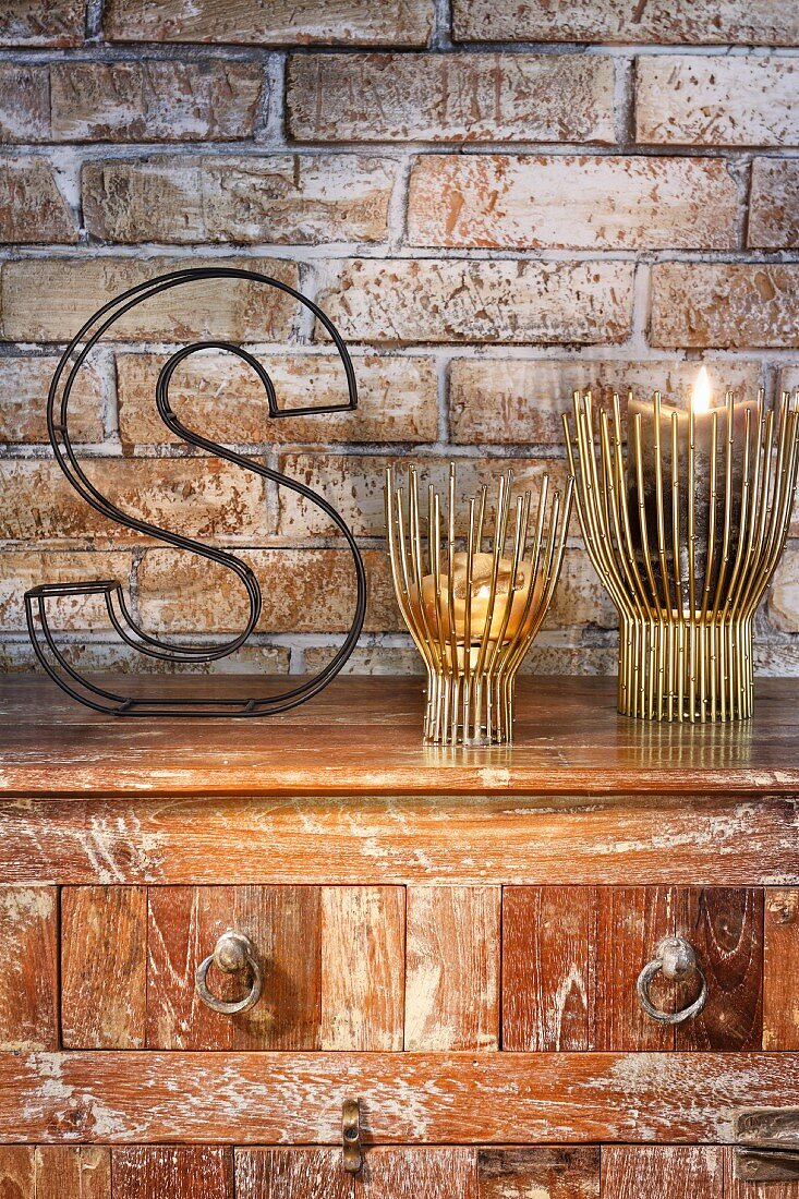 Candle lanterns and decorative wire letter on top of vintage chest of drawers against exposed brick wall