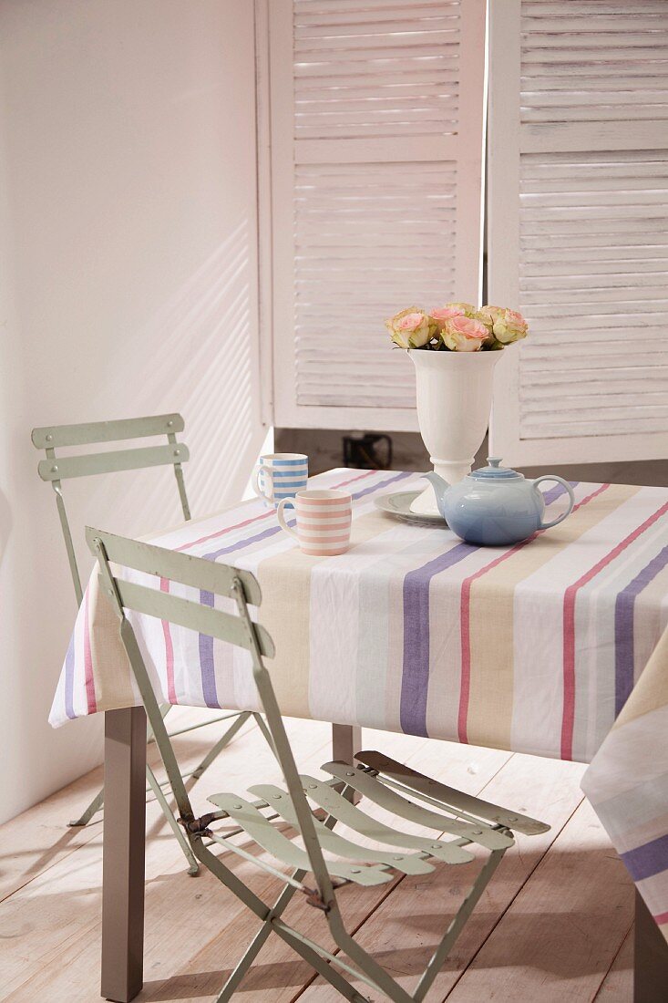 Teeservice auf Tisch mit gestreifter Tischdecke, hellgraue Klappstühle vor Fenster mit Innenläden